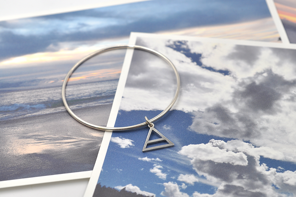 Pictured here is a round sterling silver bangle bracelet with a small air charm hanging from the continuous circle. Air symbol in a triangle with a line through the center. 