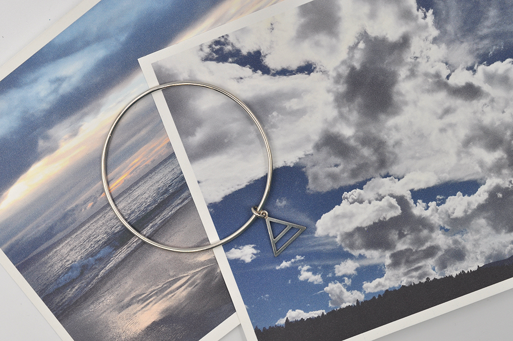 Pictured here from a birds eye view is a round sterling silver bangle bracelet with a small air charm hanging from the continuous circle. Air symbol in a triangle with a line through the center. 