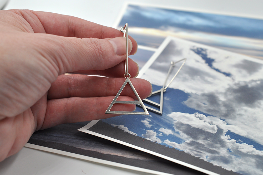 Shown here is a pair of sterling silver triangular air drop hoops in a hand for scale. These earrings showcase a large air charm hanging from the bottom point of the triangle. The air symbol featured here is a triangle with a line through the center.