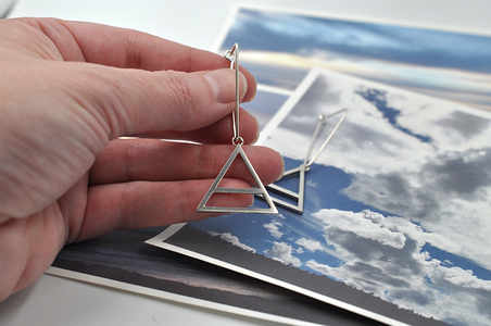 Shown here is a pair of sterling silver triangular air drop hoops in a hand for scale. These earrings showcase a large air charm hanging from the bottom point of the triangle. The air symbol featured here is a triangle with a line through the center.