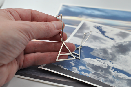 Shown here is a pair of sterling silver triangular air drop hoops in a hand for scale. These earrings showcase a large air charm hanging from the bottom point of the triangle. The air symbol featured here is a triangle with a line through the center. 