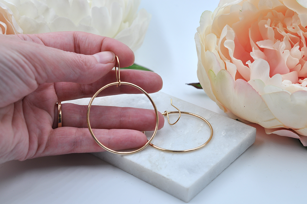 Large gold drop hoops pictured in hand for scale with another hoop earring pictured laying flat on a marble slab. 