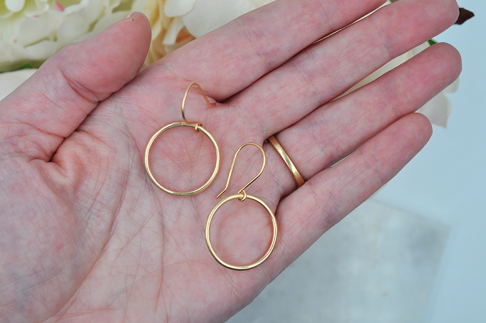 A pair of small gold drop hoops pictured in the palm of a models hand for scale.